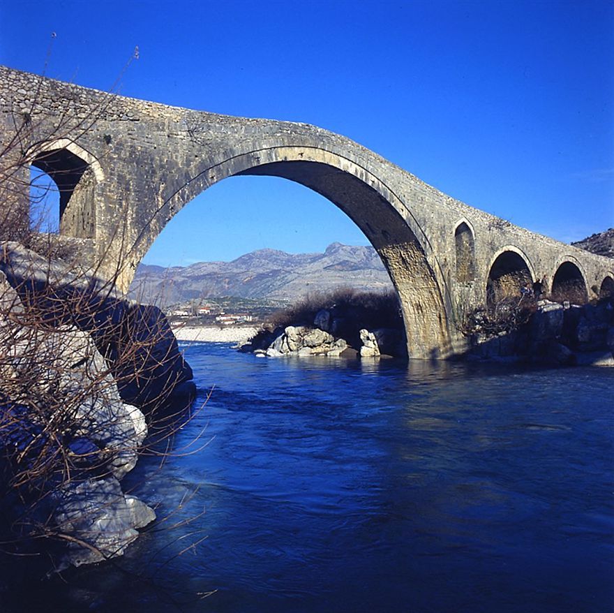 Ancient stone mystical bridge arching over a river with mountains in the background under a clear blue sky.