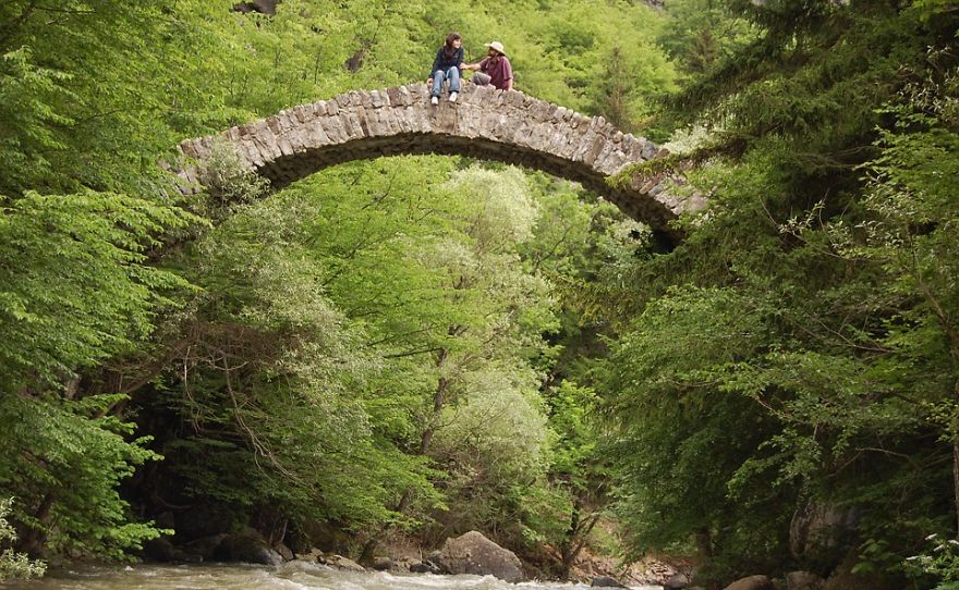 Two people sitting on a mystical stone bridge surrounded by lush green trees over a flowing river.