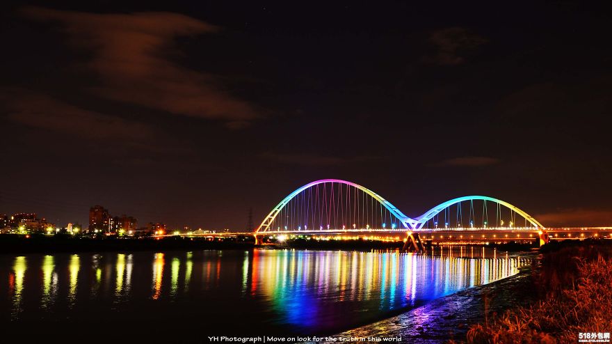 Colorful mystical bridge illuminated at night reflecting vibrant lights over calm water under a dark sky.