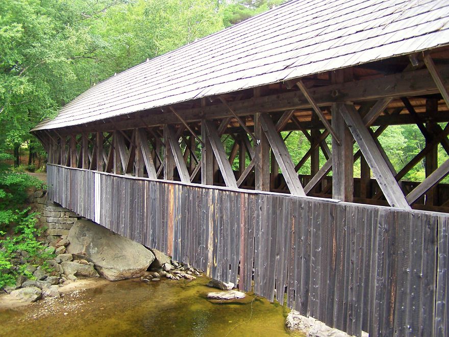 Wooden covered bridge over a shallow stream surrounded by lush green trees, a mystical bridge in a natural setting.