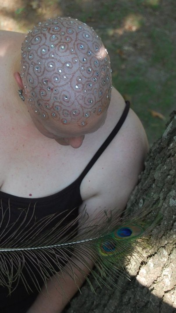 Bald head with intricate tattoo and jeweled pattern, showcasing one of the craziest haircuts ever, next to a peacock feather.