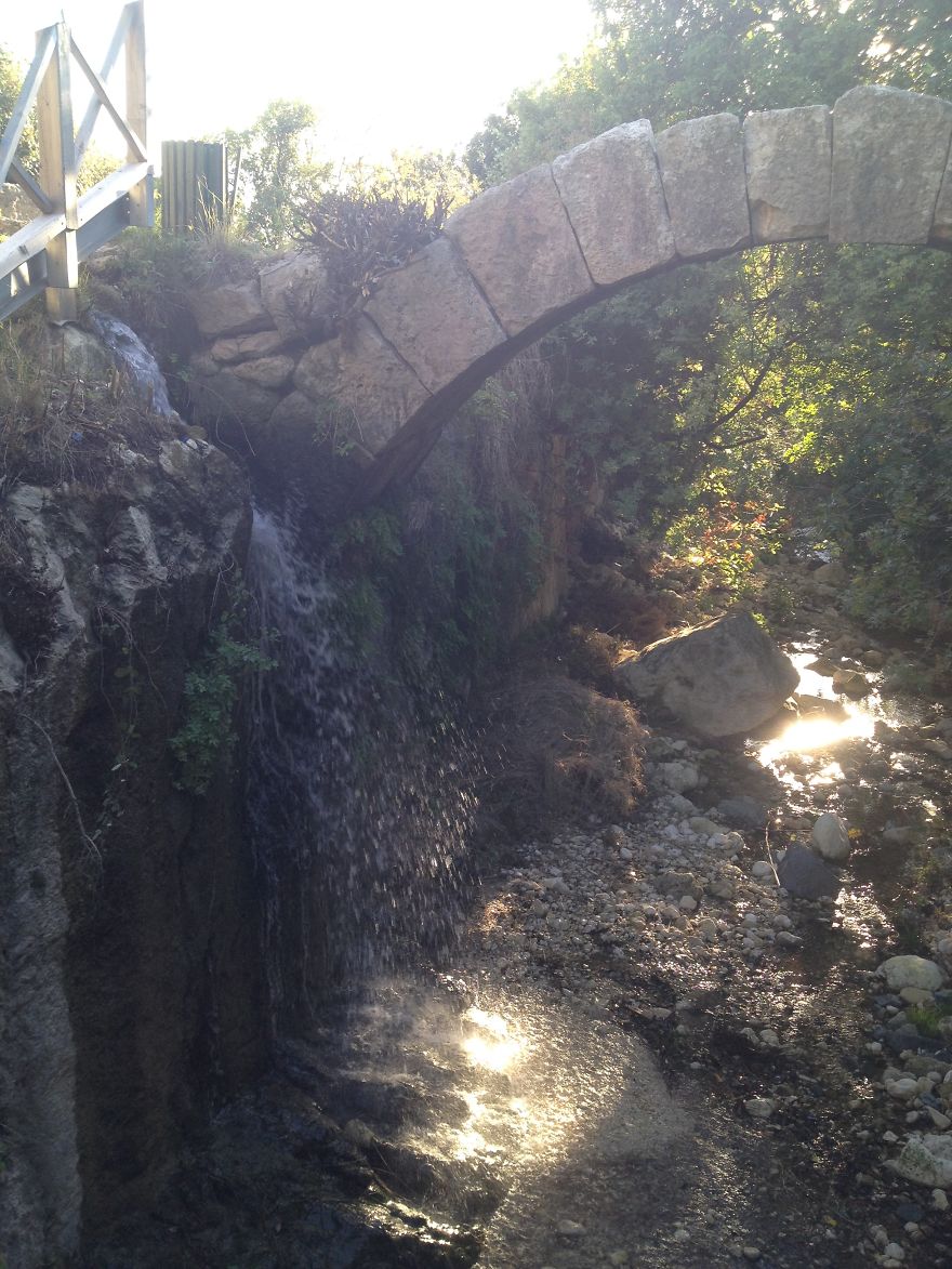 Stone mystical bridge over a small waterfall and rocky stream surrounded by sunlit trees in a natural setting
