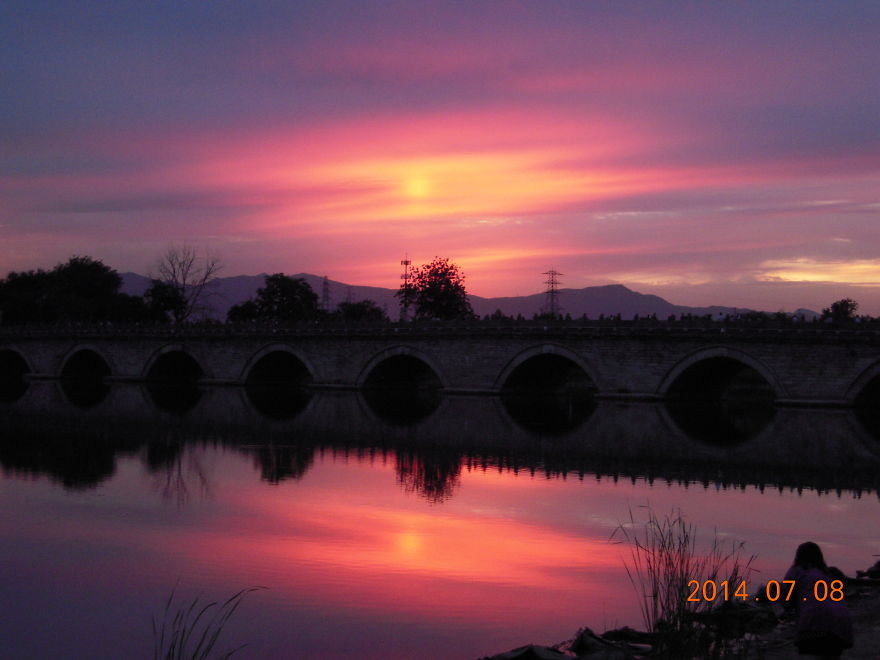 Stone bridge with arched openings reflecting in calm water at vibrant pink and purple sunset sky.