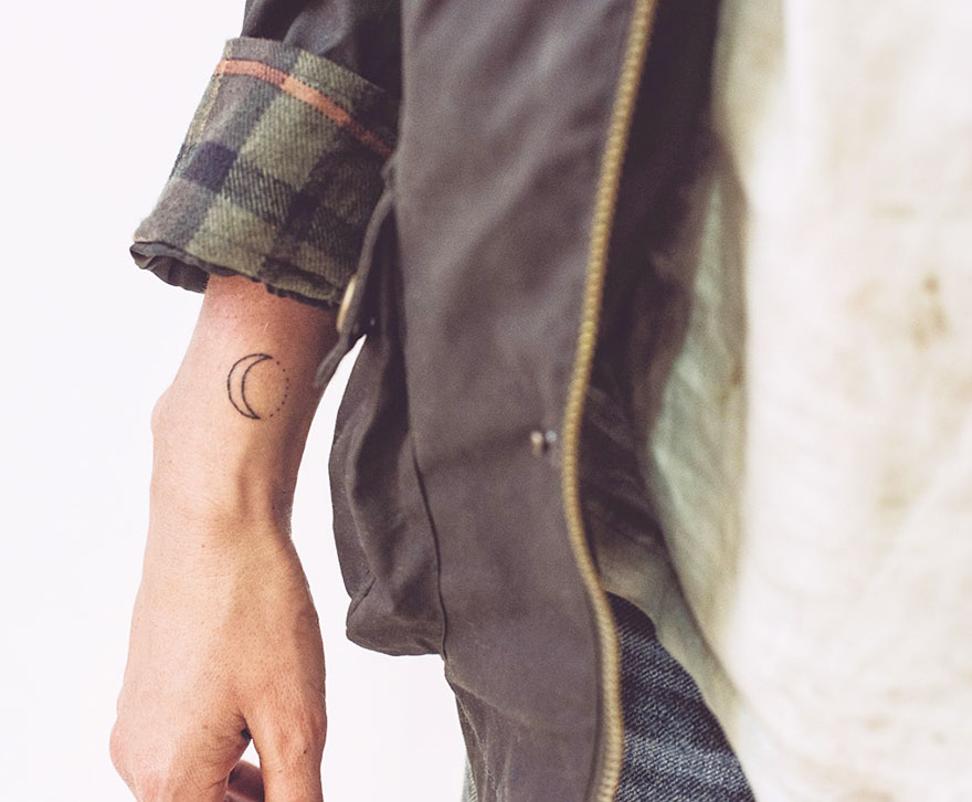 Minimalist tattoo on an arm featuring a small crescent moon, captured in a casual setting with a flannel sleeve.