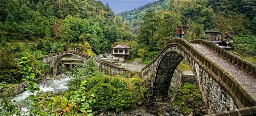 Stone mystical bridges arching over a flowing river surrounded by lush green forest and mountainous landscape.