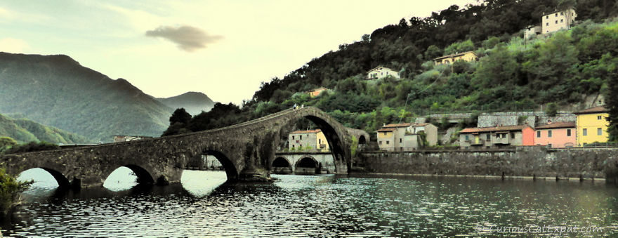 Ancient mystical stone bridge with arches crossing calm water near hillside village at sunset.