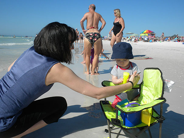 A child on a beach chair with adult nearby, beachgoers in the background, optical illusion present, causing surprise.