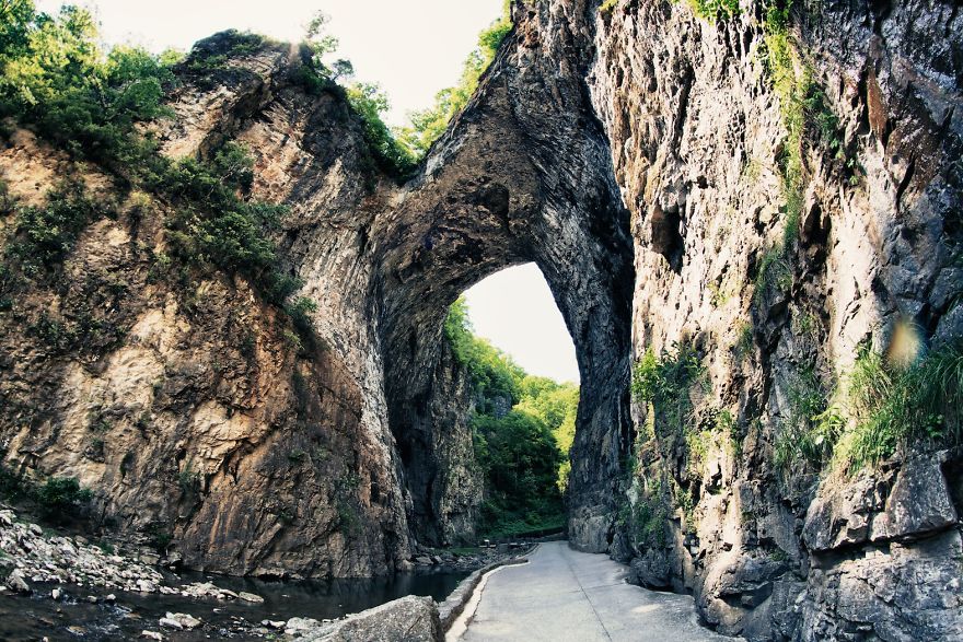 Natural rock formation creating a mystical bridge arch over a pathway surrounded by lush greenery and rugged cliffs.