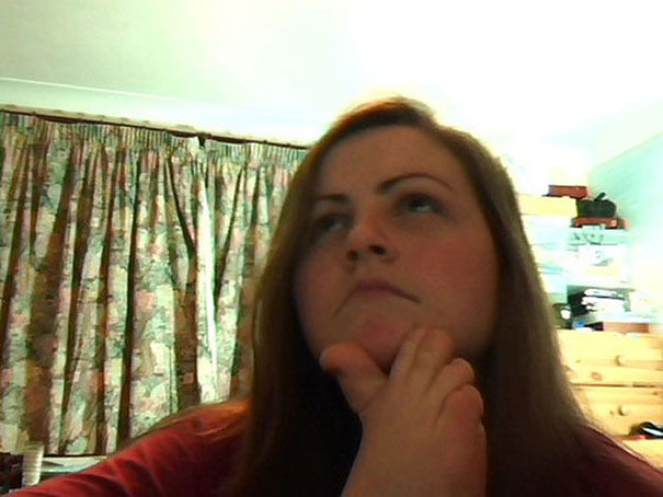 A woman sitting indoors, looking up thoughtfully, with floral curtains in the background.