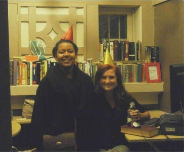 Two people wearing party hats in a dimly lit room, surrounded by books.