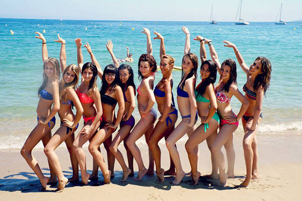 Group posing on the beach in bikinis, sailboats in the background.