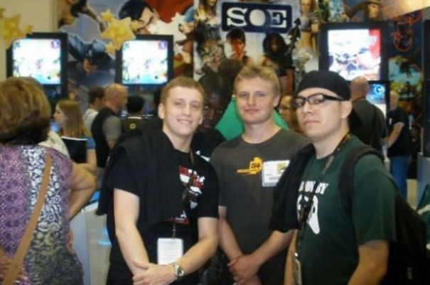 Three men pose in a crowded gaming event with vibrant posters and screens, a surprising element hidden in the scene.