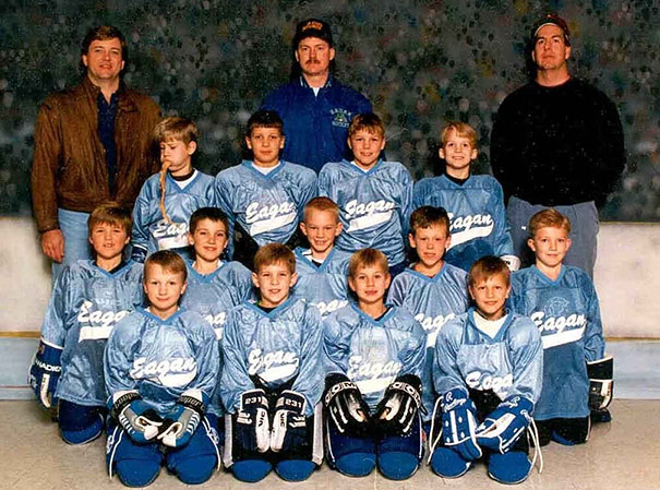 Youth hockey team group photo with three coaches, wearing blue jerseys, posed on the ice rink.