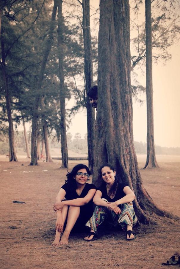 Two women sitting by a tree with a hidden person peeking, in a forest setting.