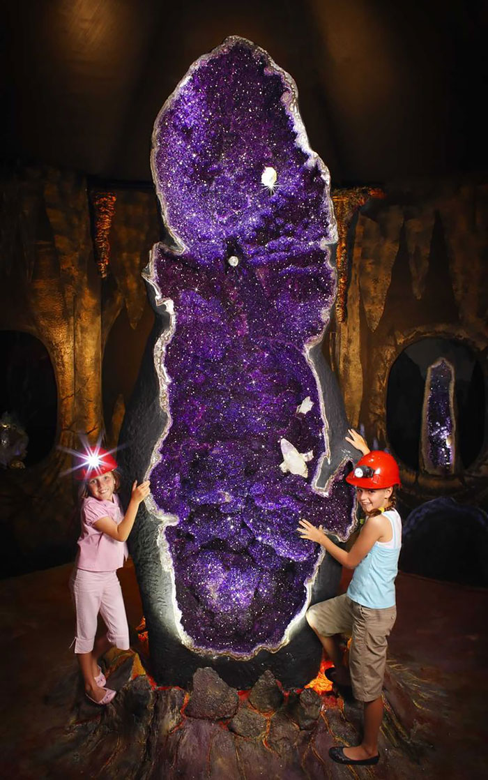 Two children with helmets admiring a large, beautiful purple mineral geode in a cave setting.