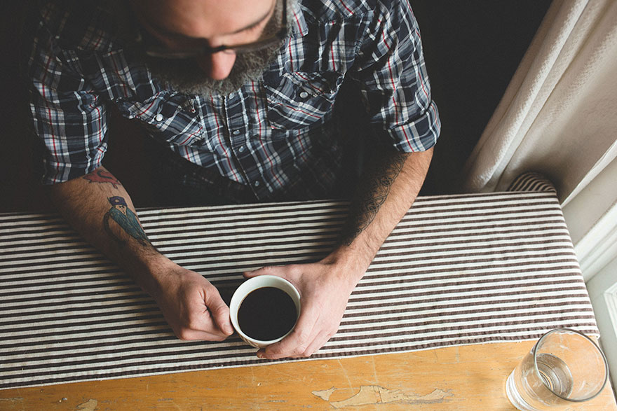 Dad's arm with son's drawing tattoos, holding coffee at a striped table.