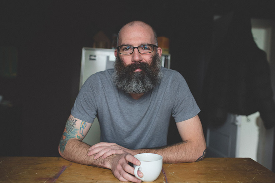 Dad with tattoos of son's drawings on arm, sitting at a table with a cup, wearing glasses and a grey t-shirt.