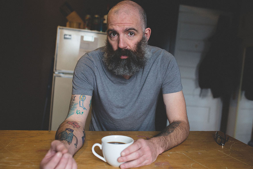 Dad with arm tattoos of son's drawings, sitting at a table with a coffee cup, wearing a gray t-shirt.