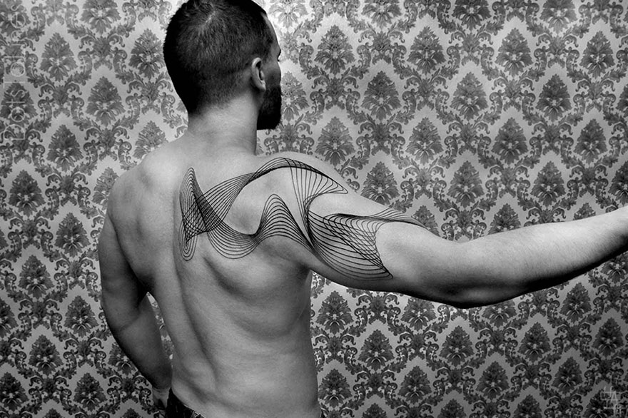 Man showcasing intricate geometric line tattoo by Chaim Machlev on back and arm against patterned wallpaper.