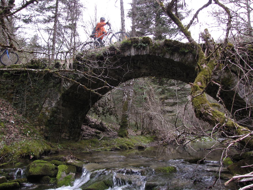 Ancient mystical bridge covered in moss over a flowing stream with a cyclist crossing in a forest setting.
