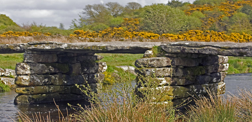 Ancient mystical stone bridge crossing a calm river surrounded by wildflowers and lush greenery in a serene landscape.