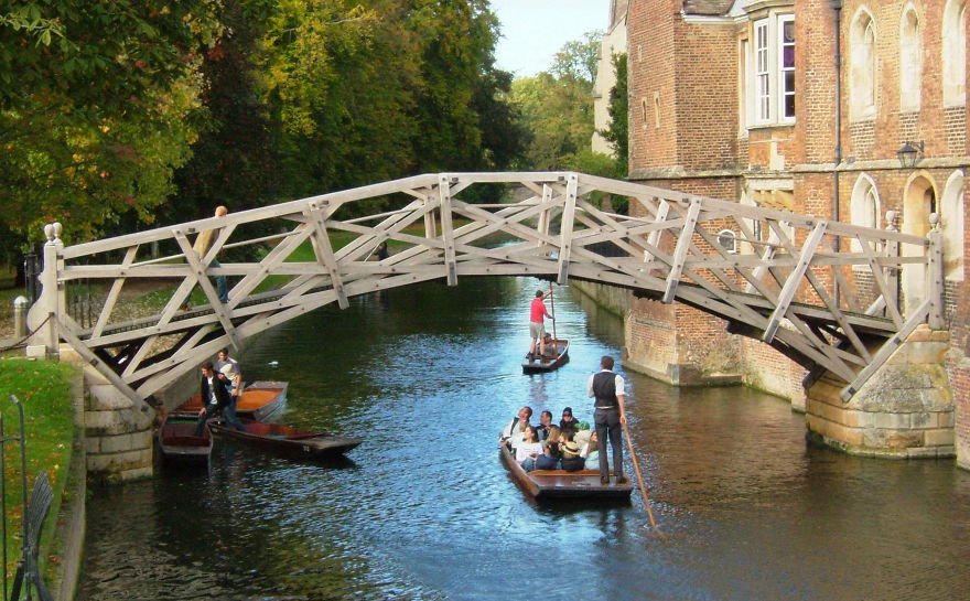 Wooden mystical bridge over a river with boats carrying people, surrounded by greenery and historic buildings.