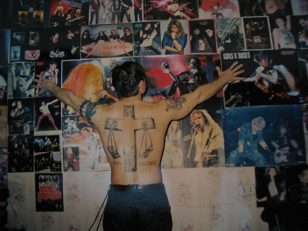 Senior with large back tattoo poses in front of rock band posters, showcasing ink at age 60.