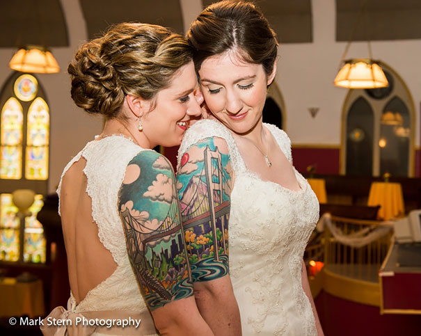 Two brides in white dresses showing matching couple tattoos featuring a colorful bridge design.