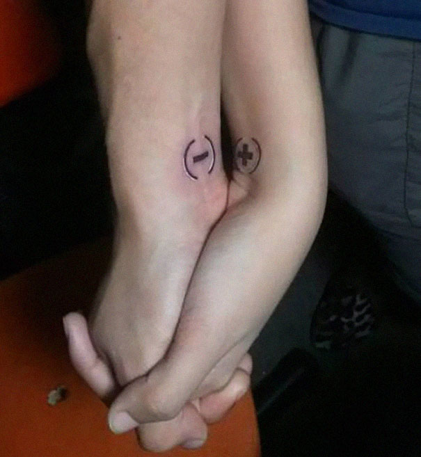 Couple holding hands, displaying matching tattoos of a plus and minus sign on their wrists.