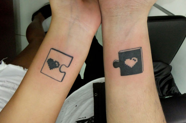 Matching couple tattoos featuring black puzzle pieces with heart shapes on wrists.