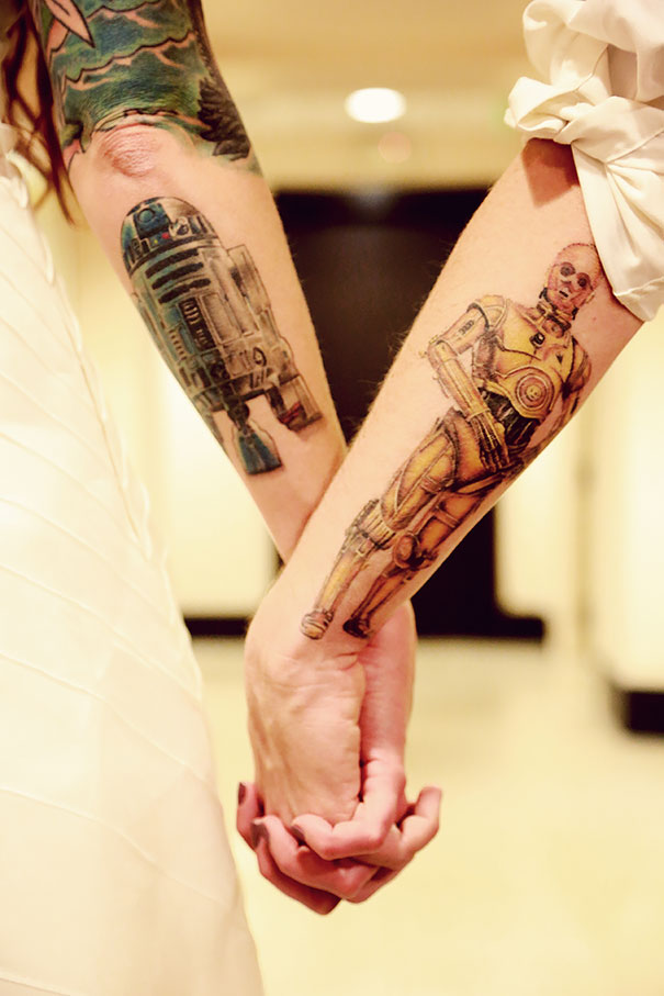 Couple holding hands with matching tattoos of R2-D2 and C-3PO on their arms celebrating love and unity.