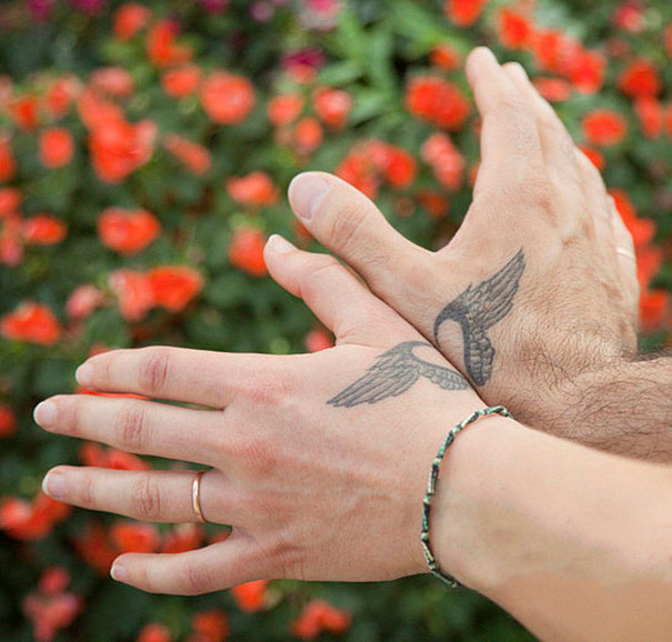 Couple's matching tattoos of wings on hands in front of vibrant flowers, symbolizing love and unity.