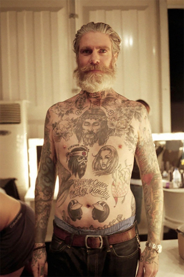 Tattooed senior with full chest and arm tattoos standing confidently in a room, showcasing ink art at age 60.