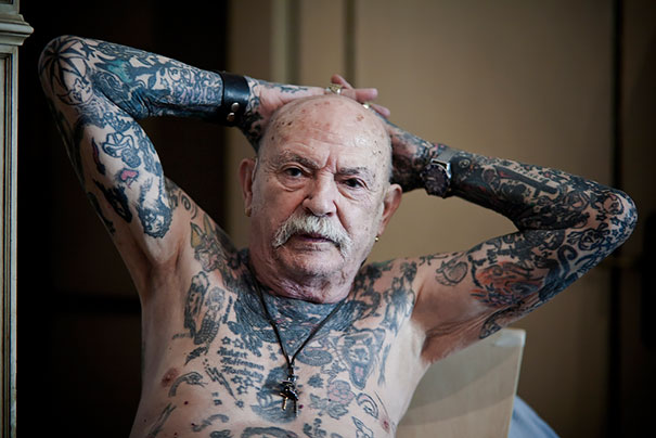 Tattooed senior with intricate body art, showing how ink looks at 60, arms resting on head, wearing wristbands and necklace.