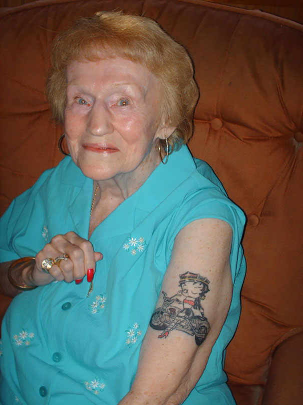 Senior woman smiling, showcasing her arm tattoo while wearing a turquoise blouse.