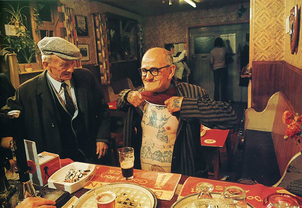 Tattooed senior in a bar showing chest tattoos to another man, illustrating how ink looks at 60.
