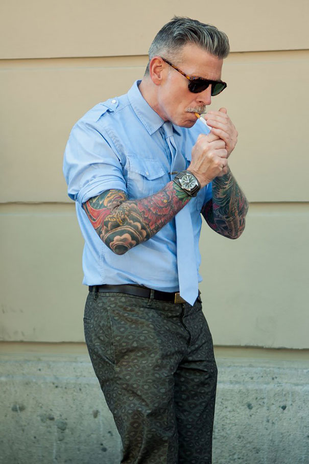 Senior with tattoos lighting a cigarette, showcasing how ink looks at 60, wearing shades and a blue shirt.