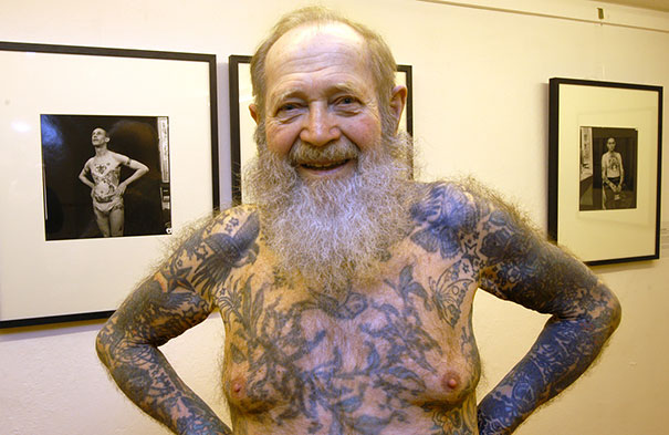 Senior with tattoos smiling, showcasing how ink looks at 60, standing in front of framed photos.