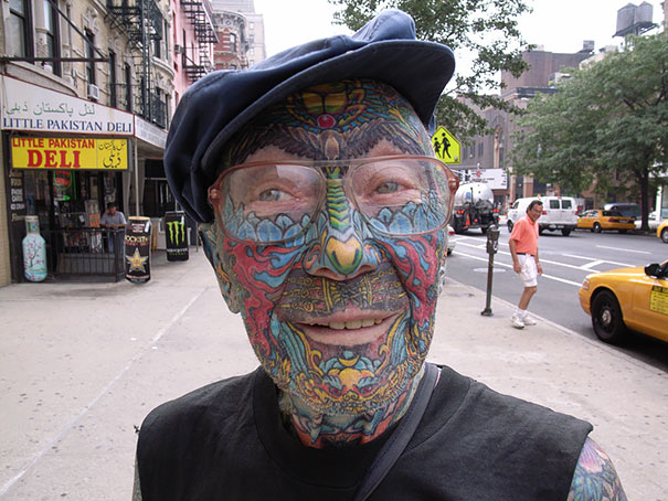Elderly man with colorful tattoos covering face and neck, wearing glasses and a cap, standing on a city street.