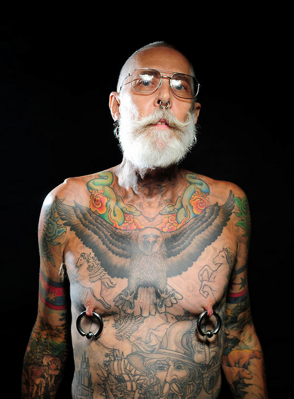 Tattooed senior with beard, showcasing extensive tattoos including an eagle on chest against a dark background.