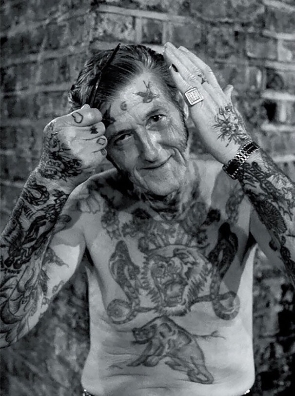 Senior man with tattoos combing hair, showcasing ink on chest and arms against a brick background.