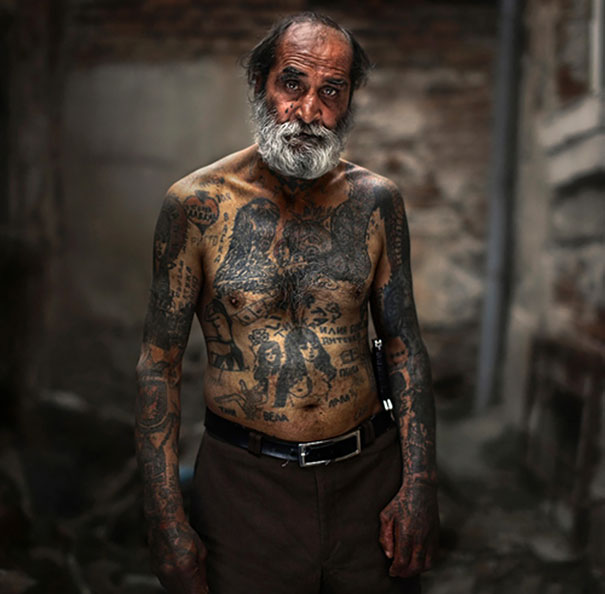 Elderly man with tattoos showing how ink looks on senior skin, standing in a rustic setting.