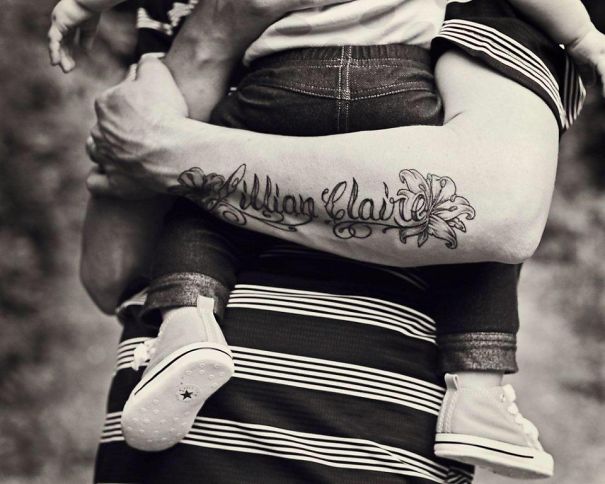 Tattooed parent holding a baby, showcasing arm tattoo with the name "Jillian Claire" in a striped shirt.