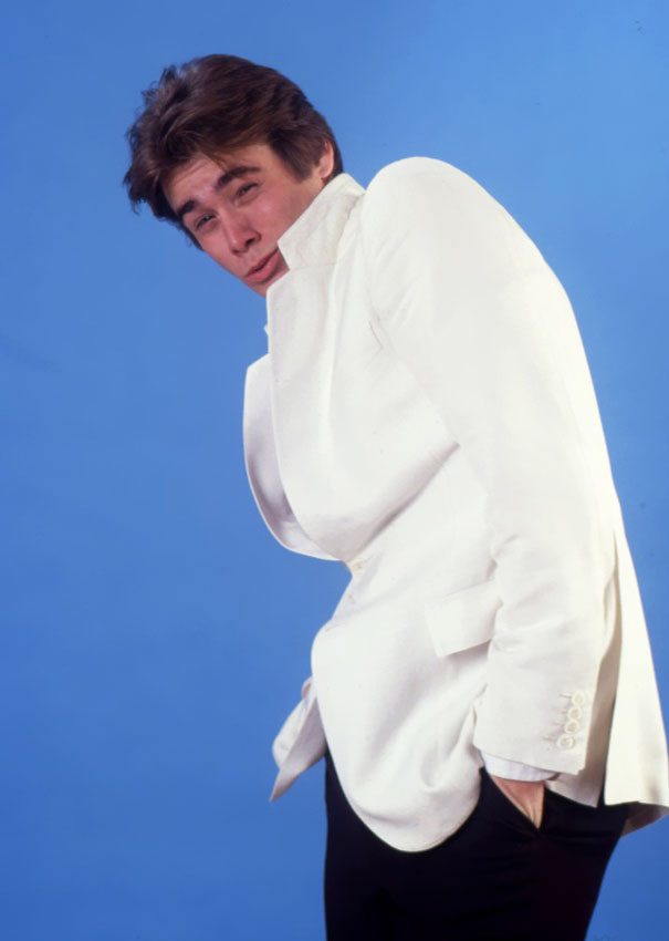 Young Jim Carrey in a white jacket posing with a playful expression against a blue background for celeb impressions.