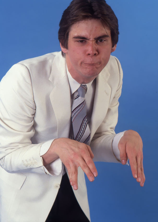 Young Jim Carrey in a white suit and patterned tie performing manic celeb impressions with expressive facial gestures. Young Jim Carrey in a white suit and patterned tie performing manic celeb impressions with expressive facial gestures.