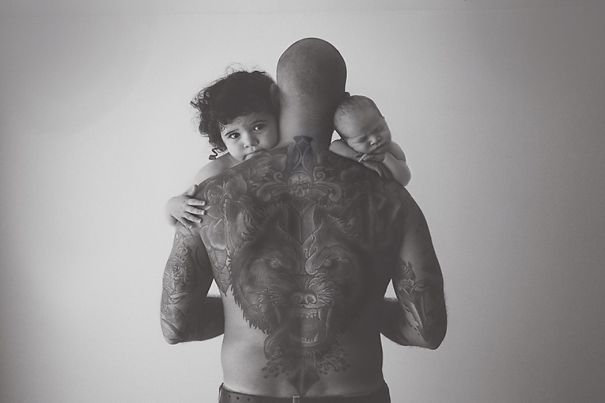 Tattooed parent holding two babies, showcasing an intricate wolf tattoo across their back.