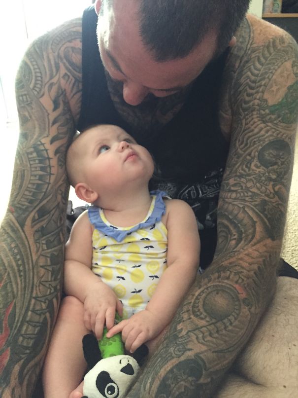 Tattooed parent holding baby who gazes up, both sharing a tender moment together.