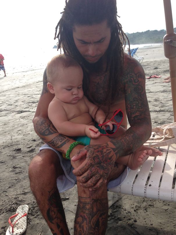 Tattooed parent holding a baby on the beach, showcasing beautiful body art and family bond.