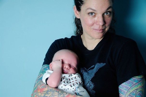 Tattooed parent holding a baby, both smiling warmly against a blue background.