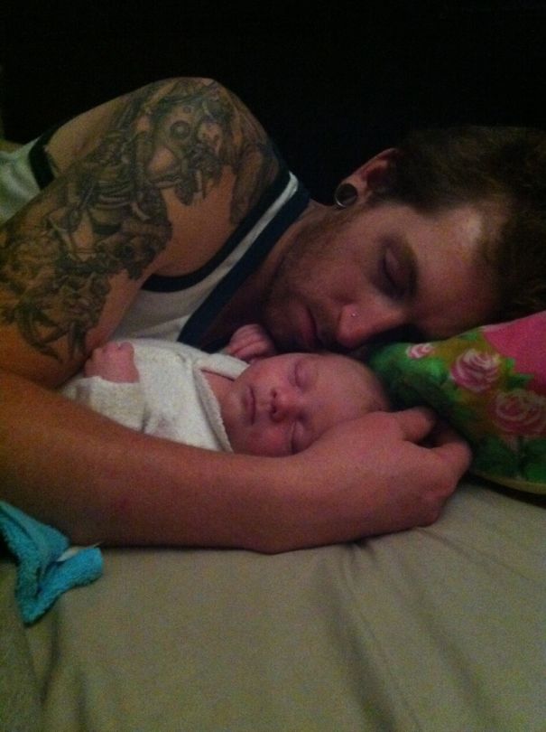 Tattooed parent peacefully sleeping beside swaddled baby on a bed, showcasing a tender moment together.